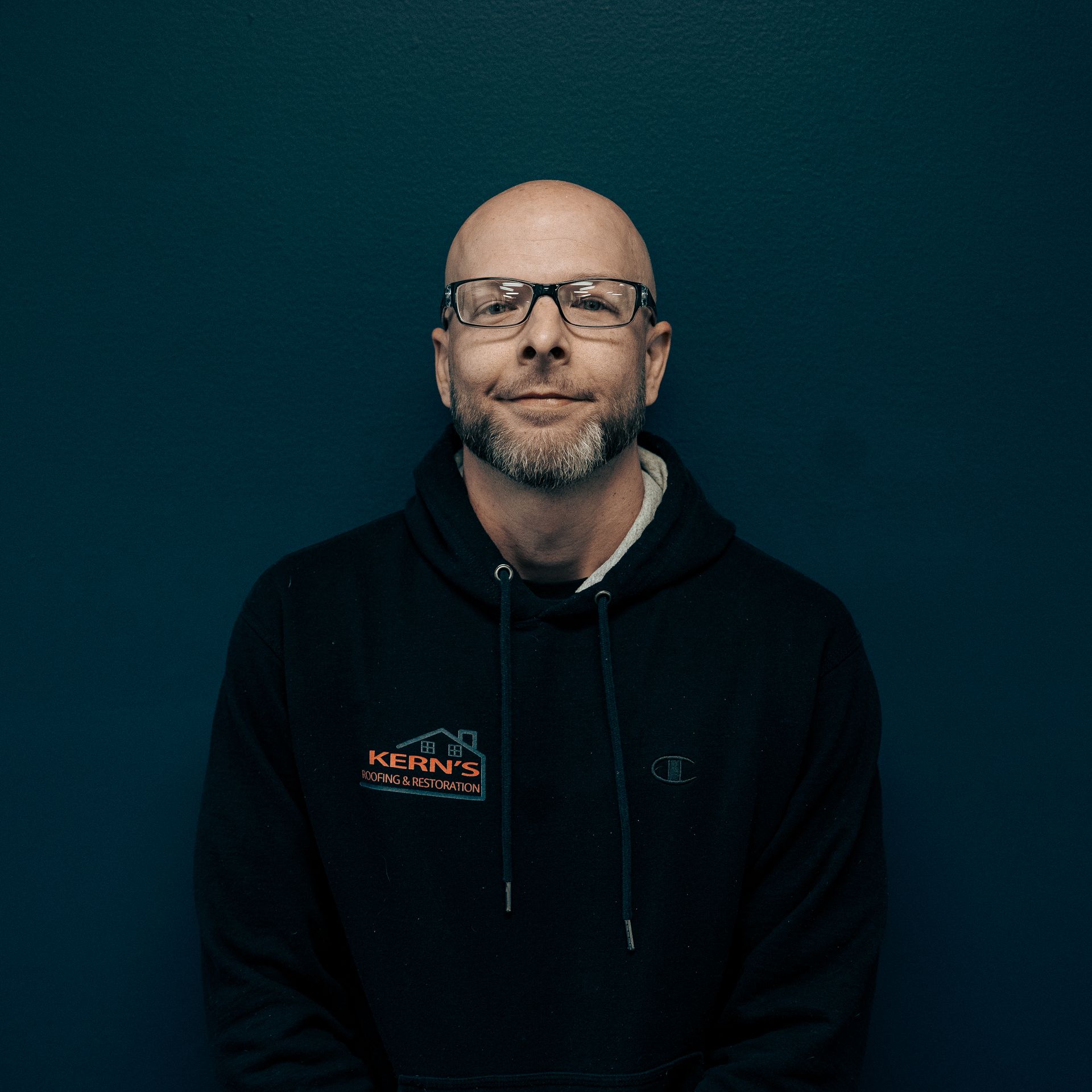 Man wearing glasses and a black hoodie, seated against a dark blue background.
