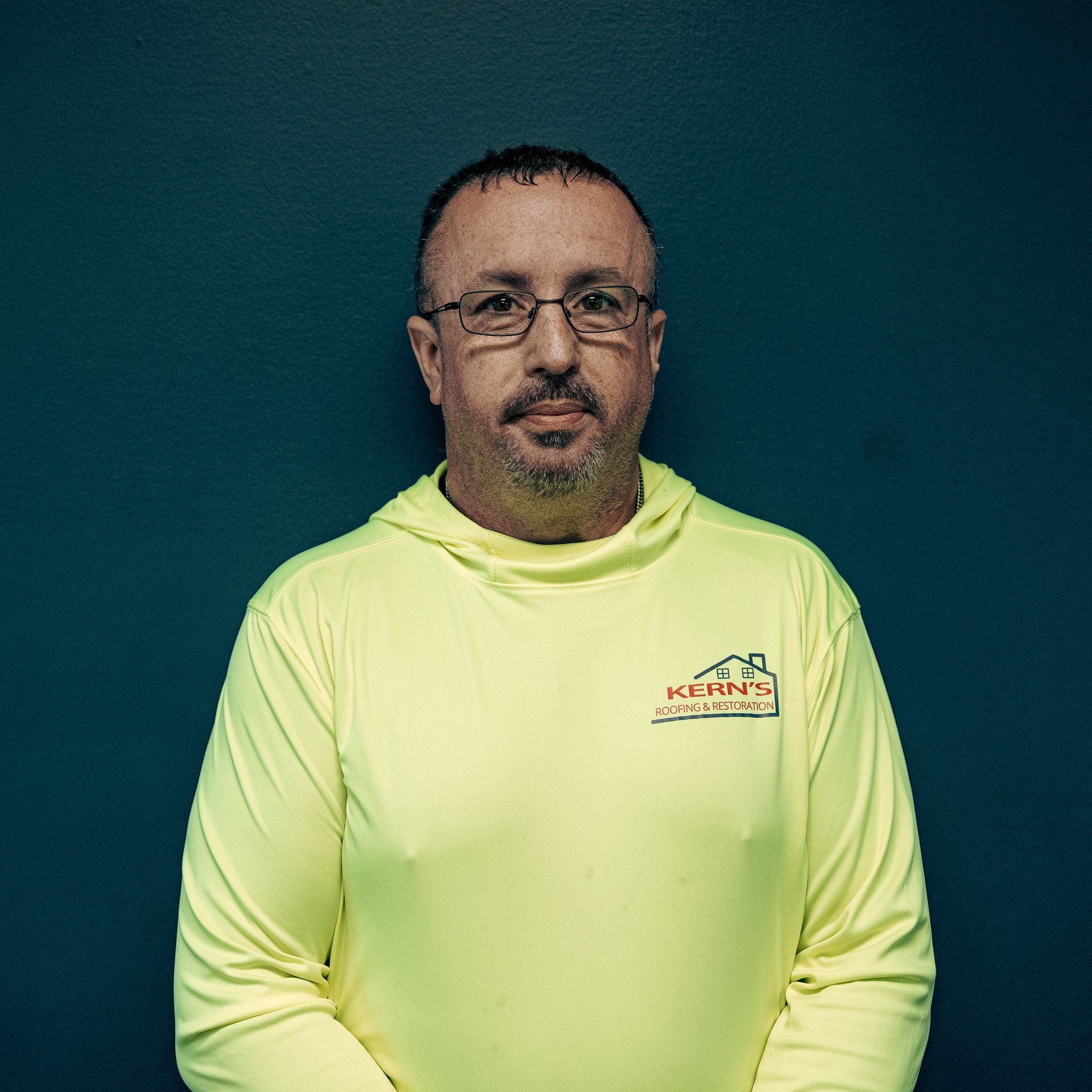 Man standing against a teal background, wearing a neon yellow shirt with a small logo