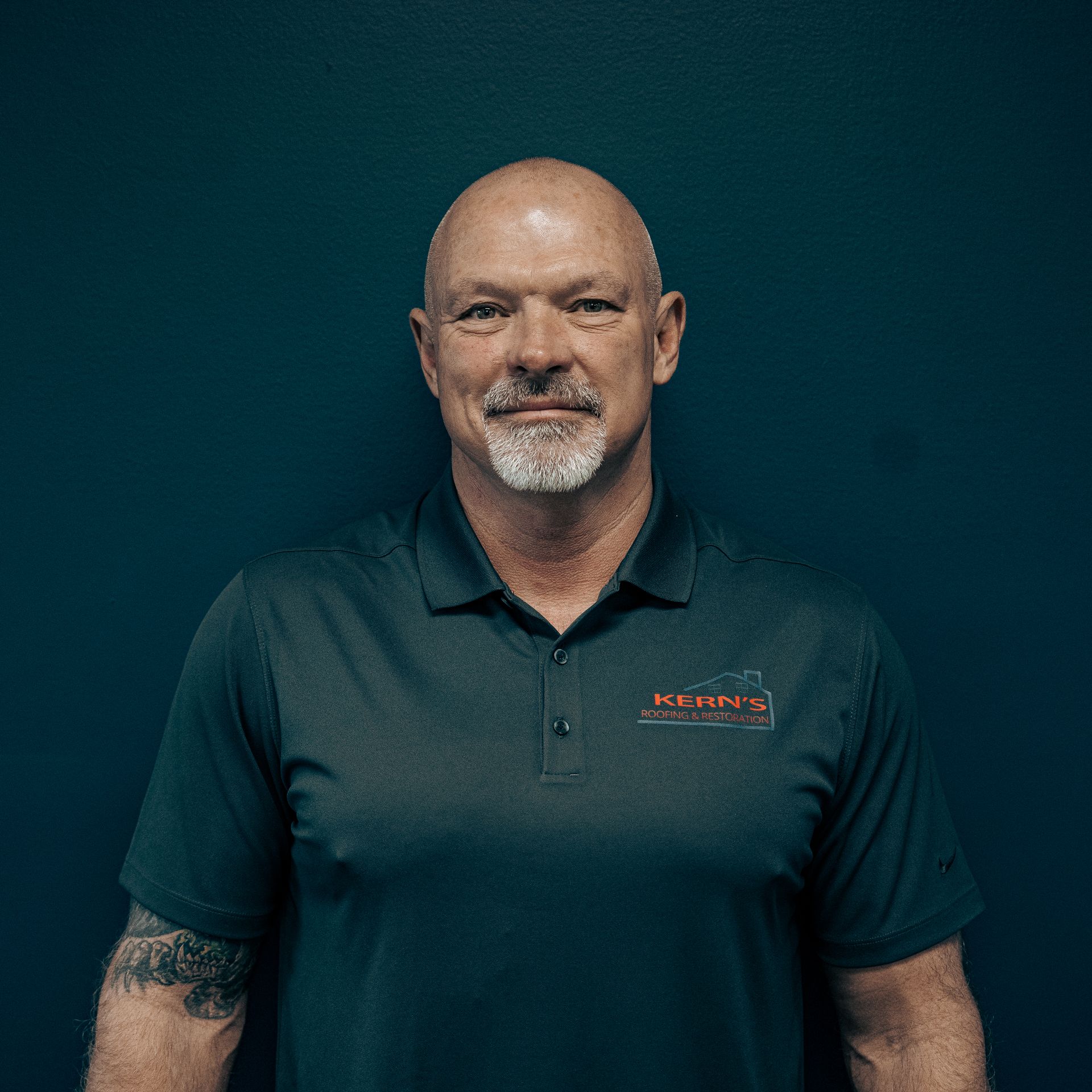 Portrait of a bald man with a goatee wearing a dark polo shirt against a teal background