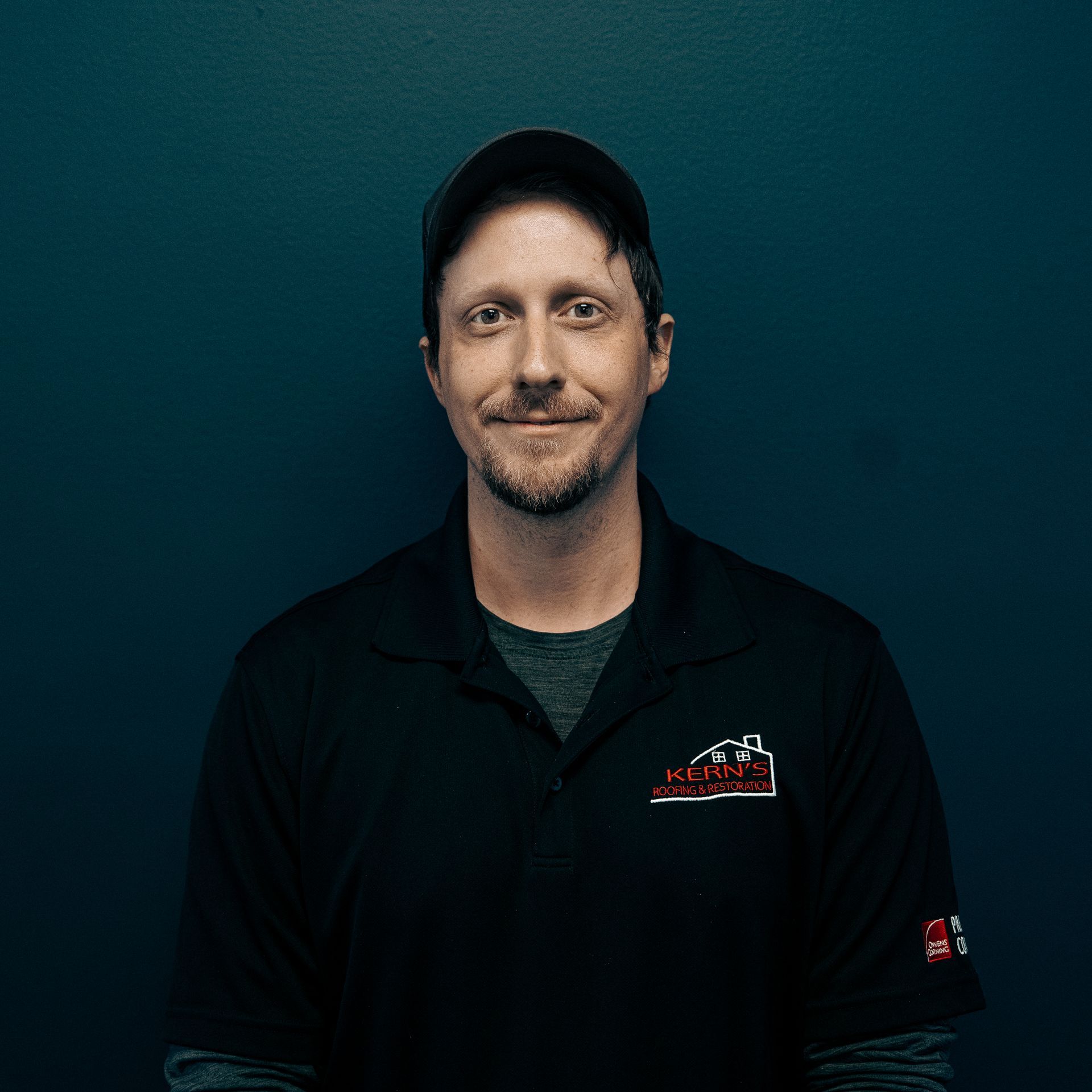 Smiling man in black cap and shirt against a dark teal background.