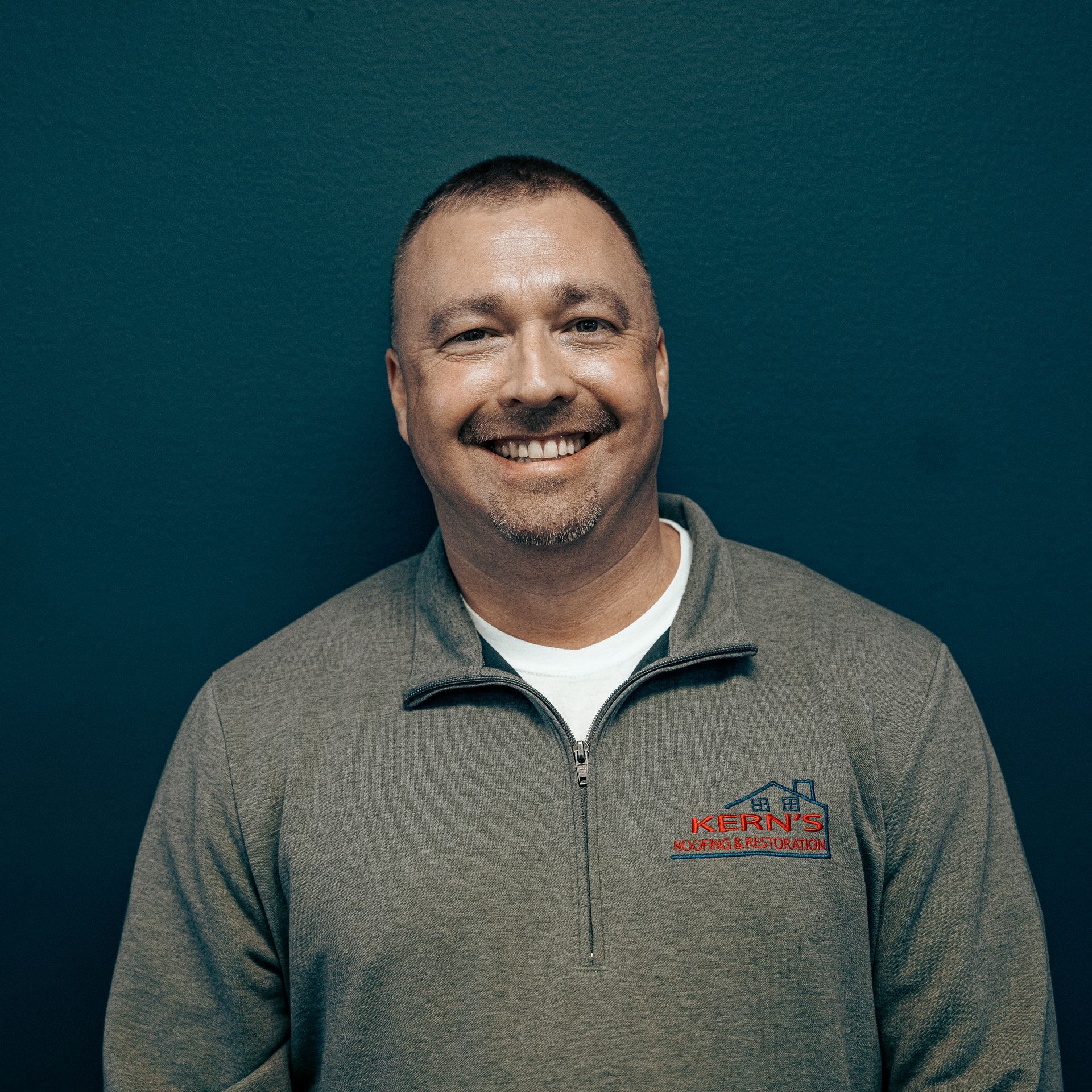 Smiling man in a gray quarter-zip pullover against a teal background