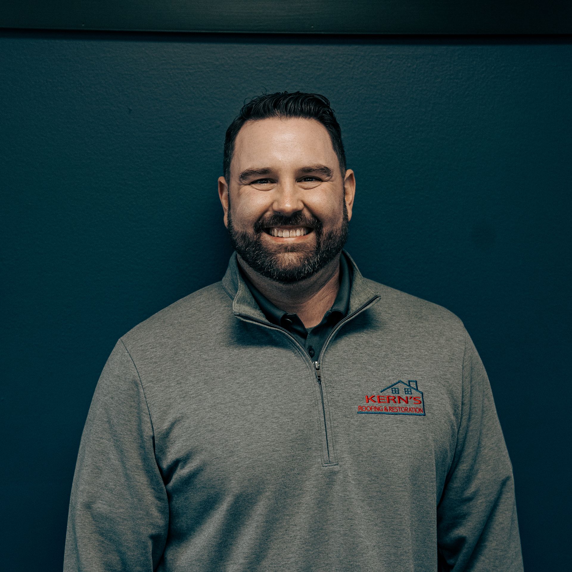 Smiling man in a gray quarter-zip sweater against a dark teal background
