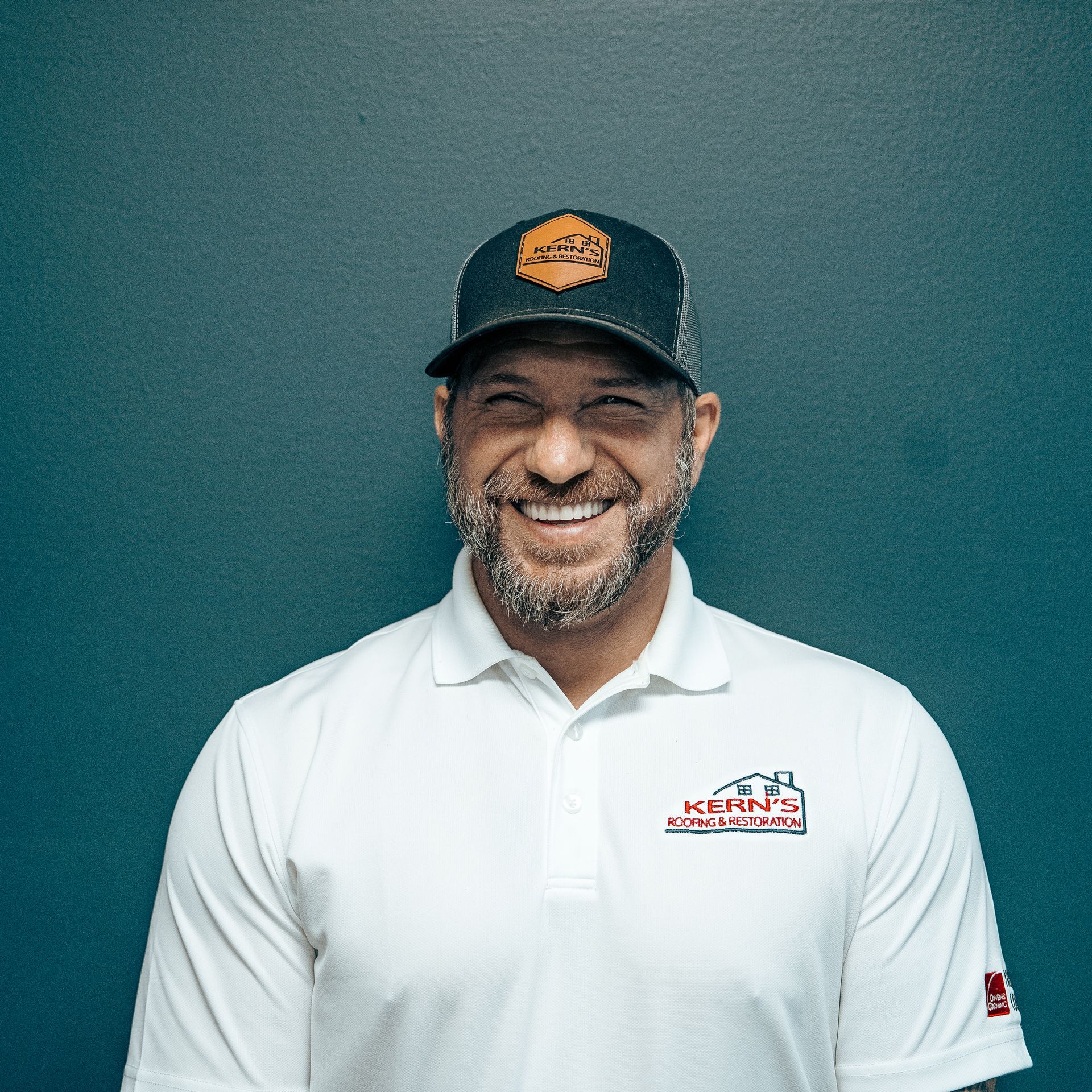 Smiling man in a white polo and black cap against a teal wall