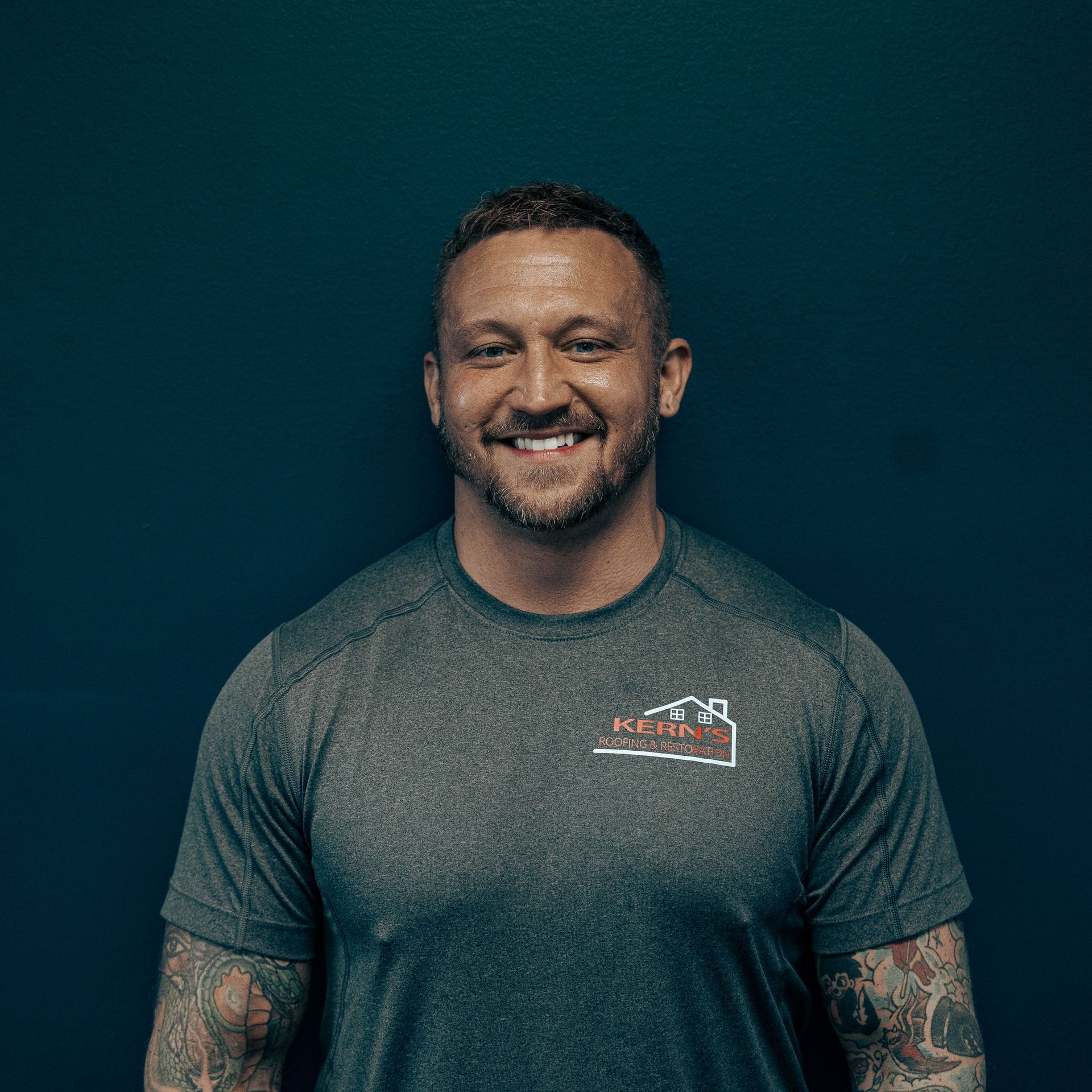 Smiling man with tattooed arms in a gray T-shirt against a dark teal background