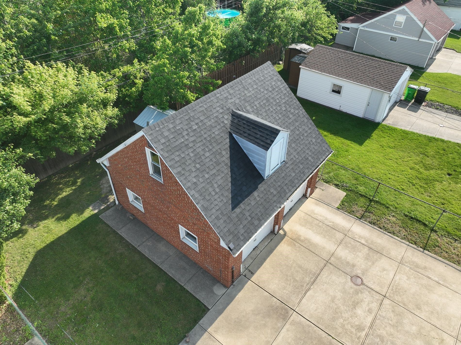 Aerial view of a two-story brick house with a dark gray roof and a gray concrete driveway. Trees surround the property.