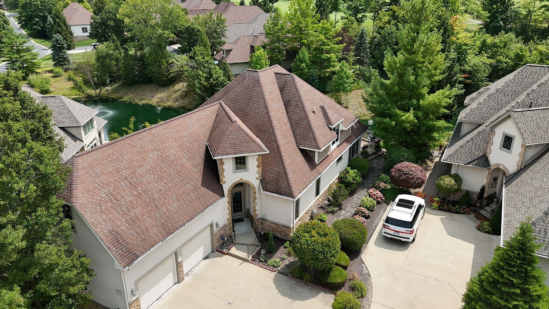 Aerial view of a luxury home with a brown roof, white exterior, and three-car garage. A white SUV sits in the driveway.
