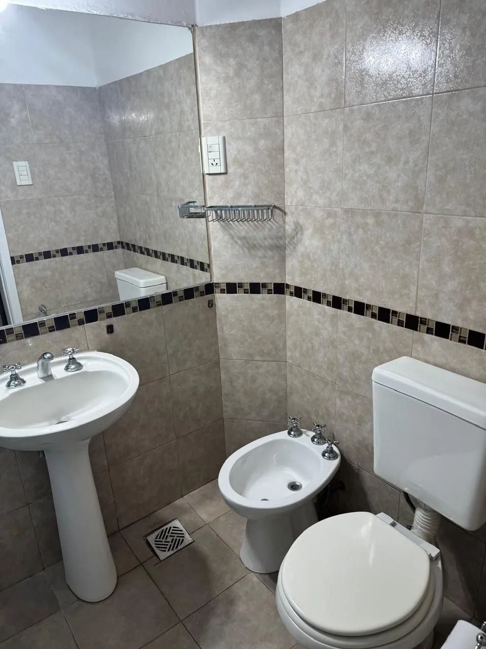 Un baño con lavabo de pedestal, bidé e inodoro, con paredes revestidas de azulejos beige