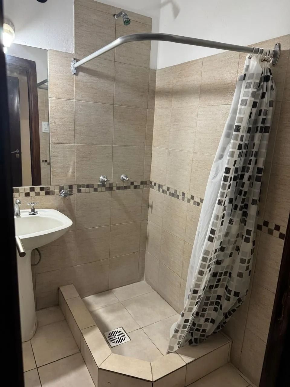 Un baño que muestra una ducha a ras de suelo con azulejos, una cortina estampada y un lavabo blanco con pedestal adyacente.