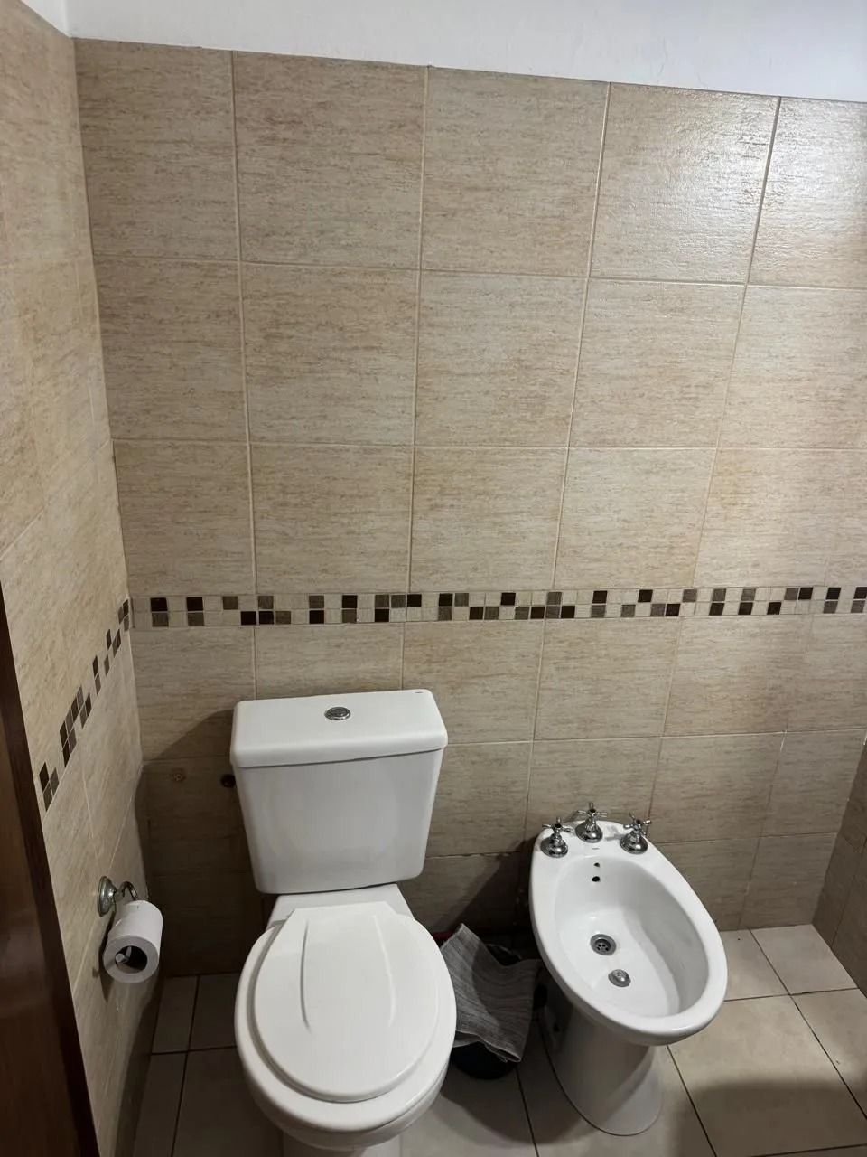 Un inodoro y un bidé blancos sobre una pared de azulejos beige con un borde decorativo de mosaico marrón.