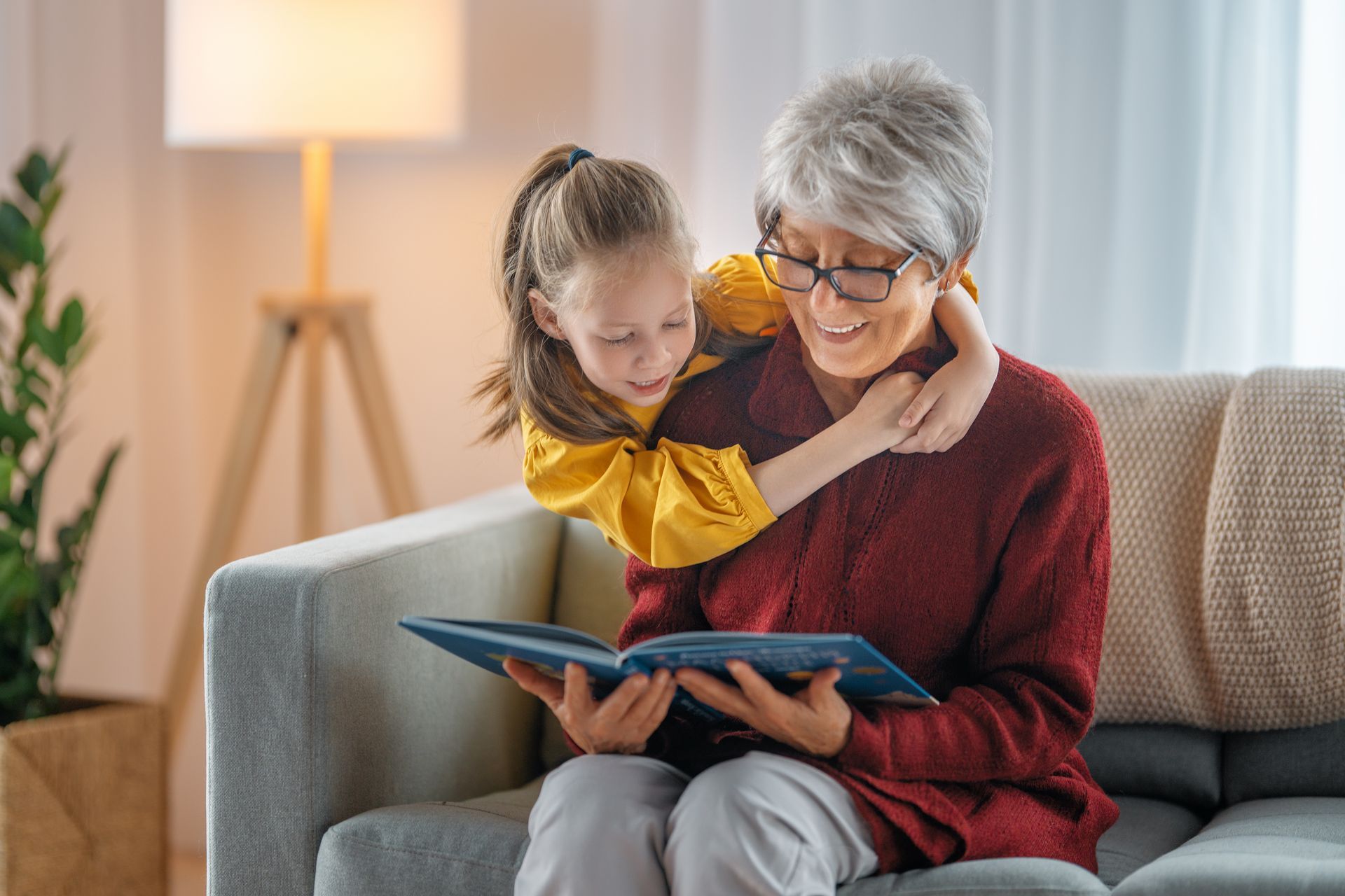 Girl hugs woman reading a book on a couch indoors.