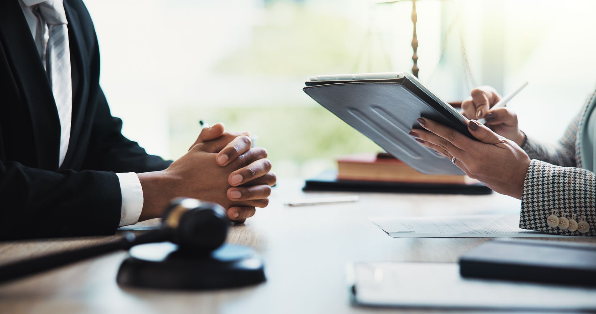 Man in suit sits at table, listens as person signs document. Gavel and scale of justice visible.