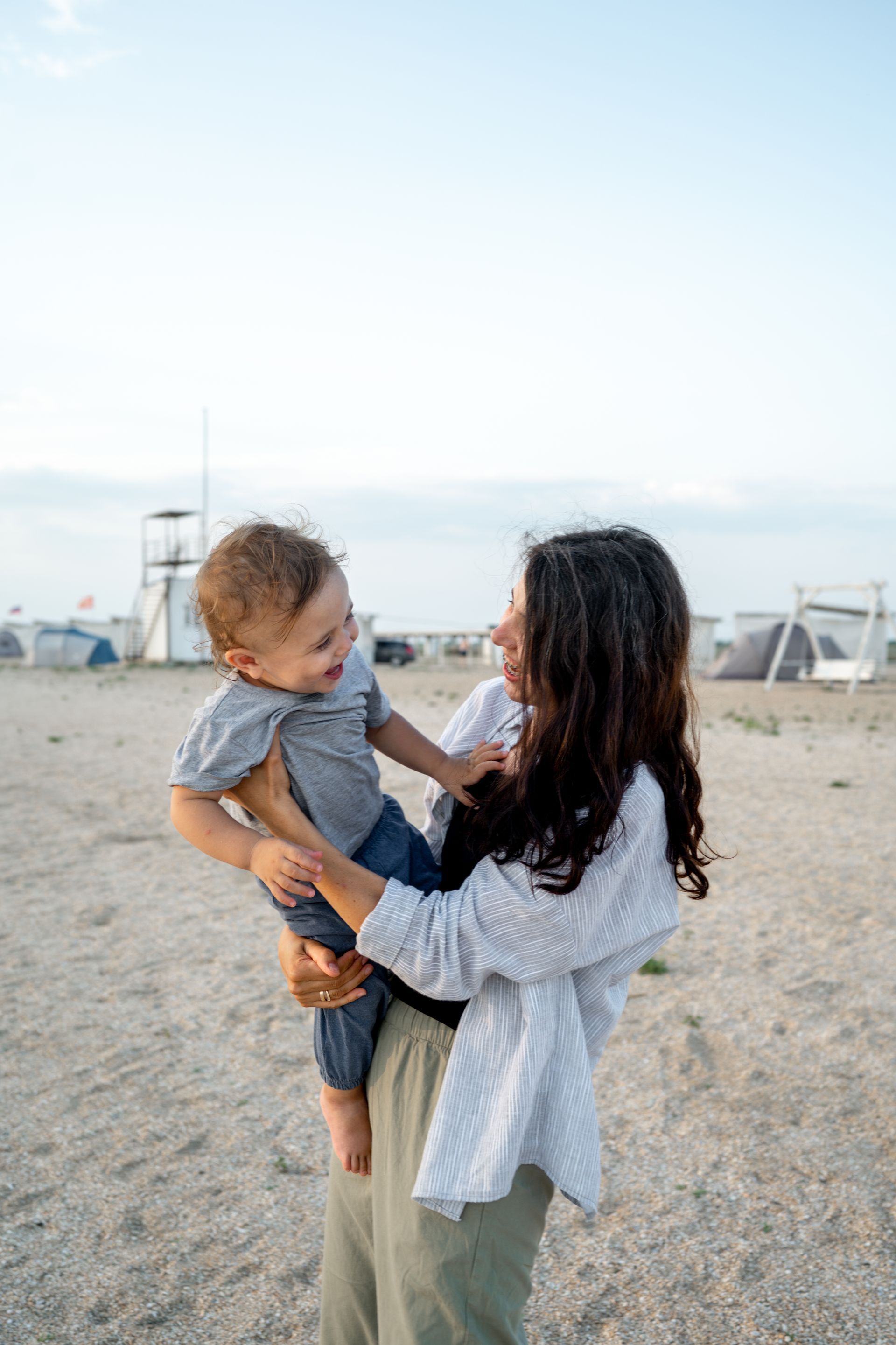 Woman holding a child on a sandy beach, both smiling. Blue sky backdrop.