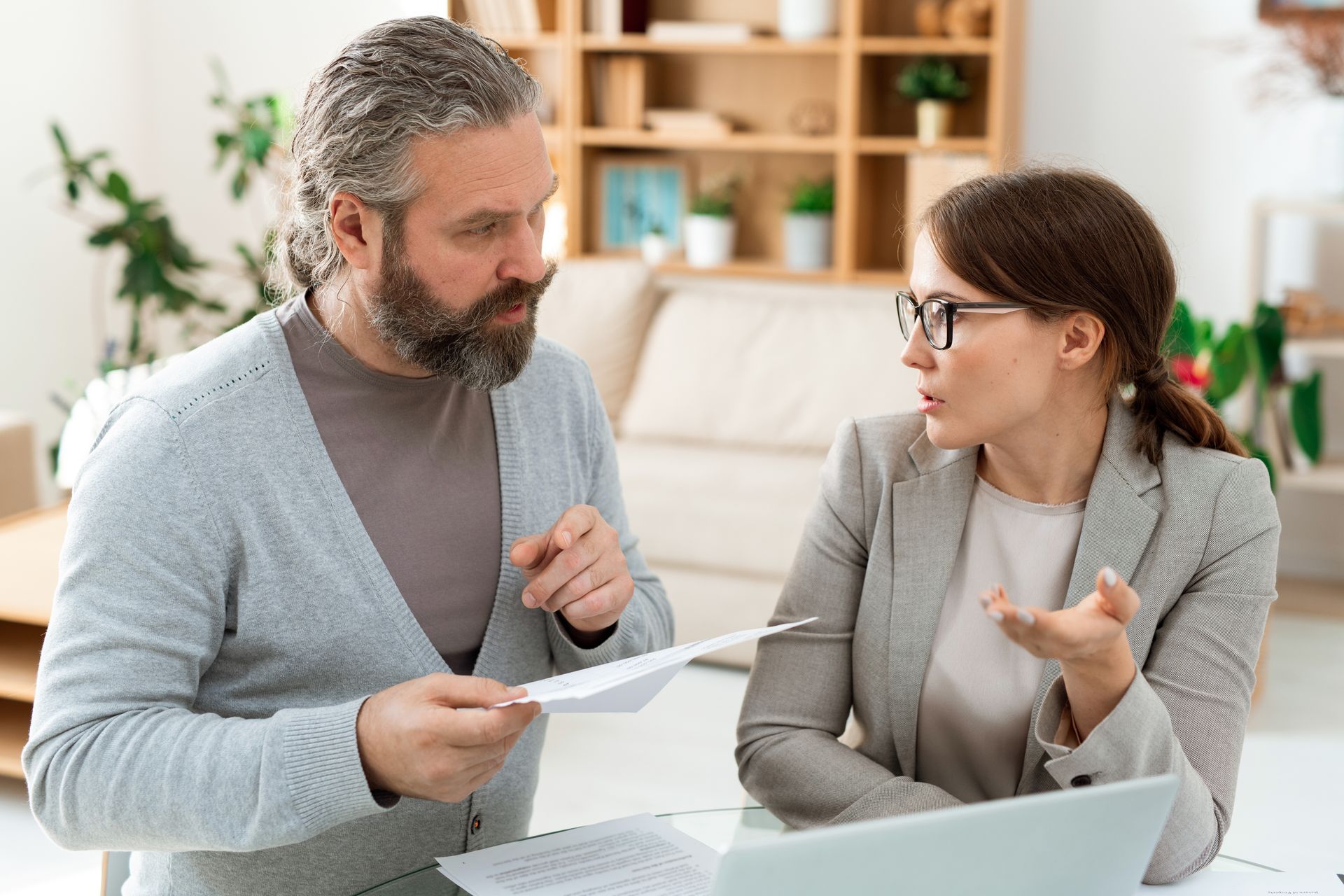 A man and woman review paperwork at a table with a laptop. The woman gestures, and the man holds papers.
