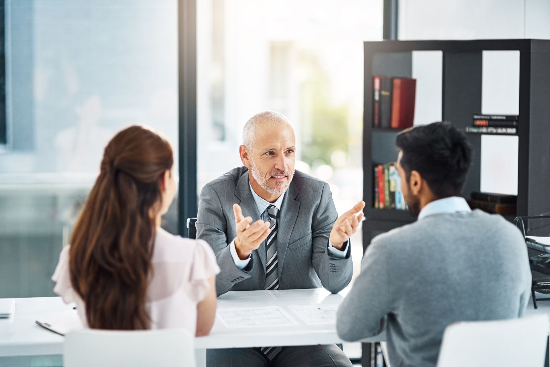 Man in suit gestures as he speaks to a couple seated at a table in an office setting.