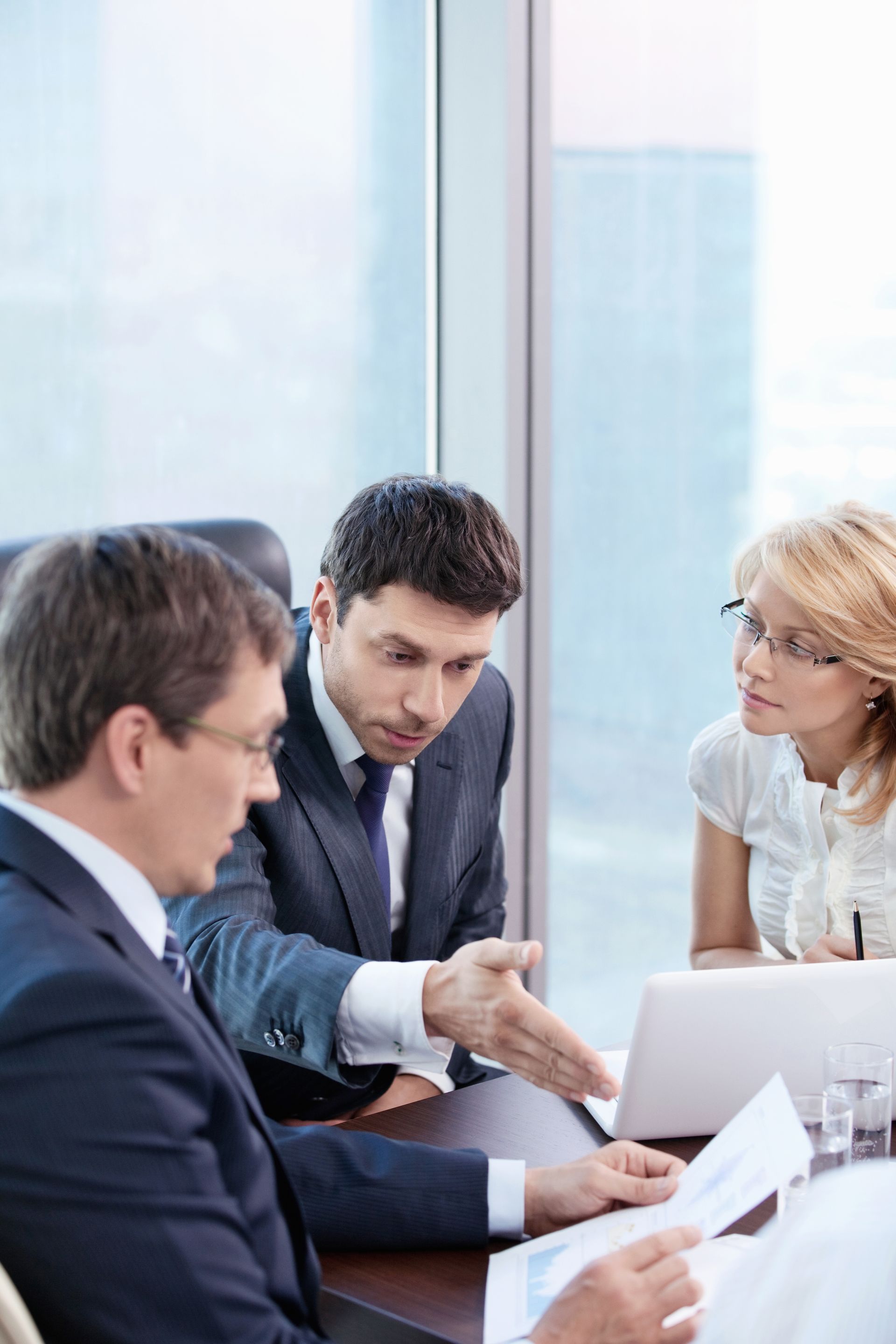 Three people in suits at a conference table, reviewing documents, one gesturing.