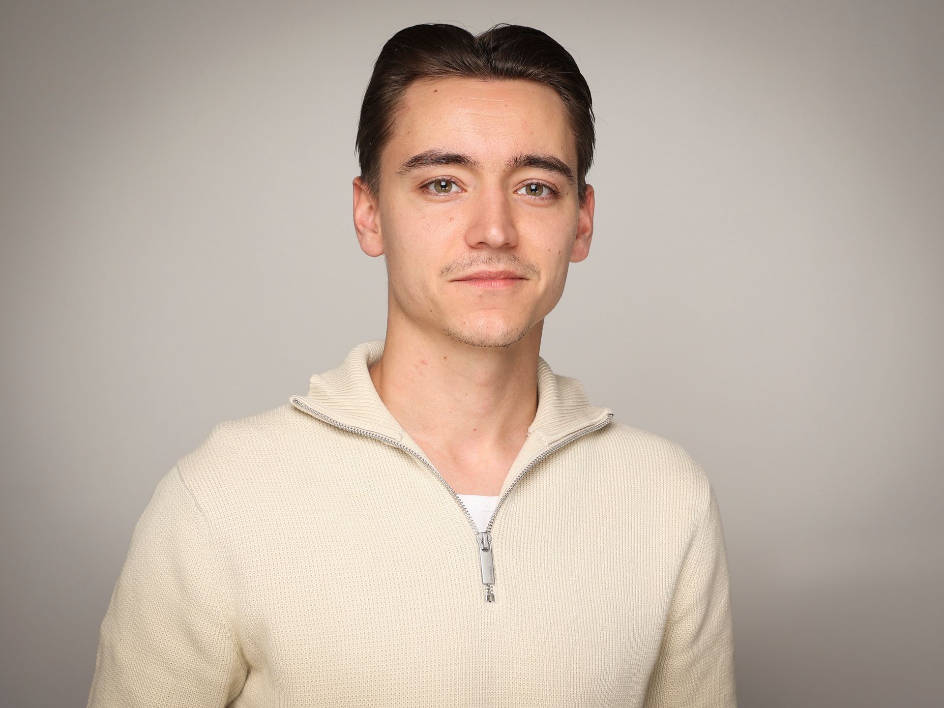 Ein junger Mann in einem weißen Pullover steht vor einem grauen Hintergrund.
