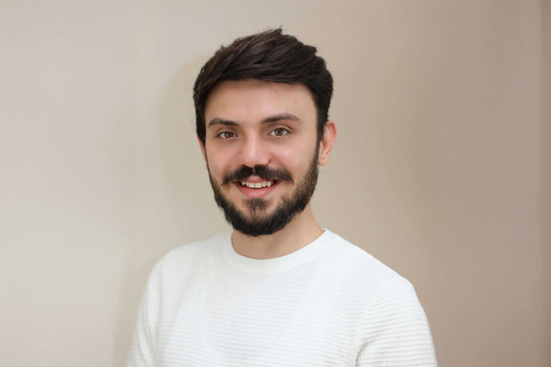 Lächelnder Mann mit dunklem Haar und Bart, der einen weißen Pullover trägt und an einer hellbeigen Wand lehnt.