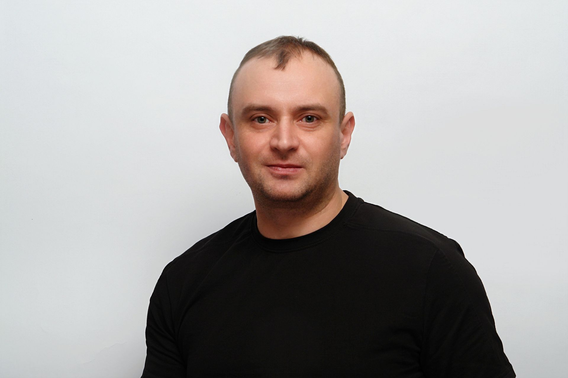 Ein Mann in einem schwarzen Hemd steht vor einer weißen Wand.