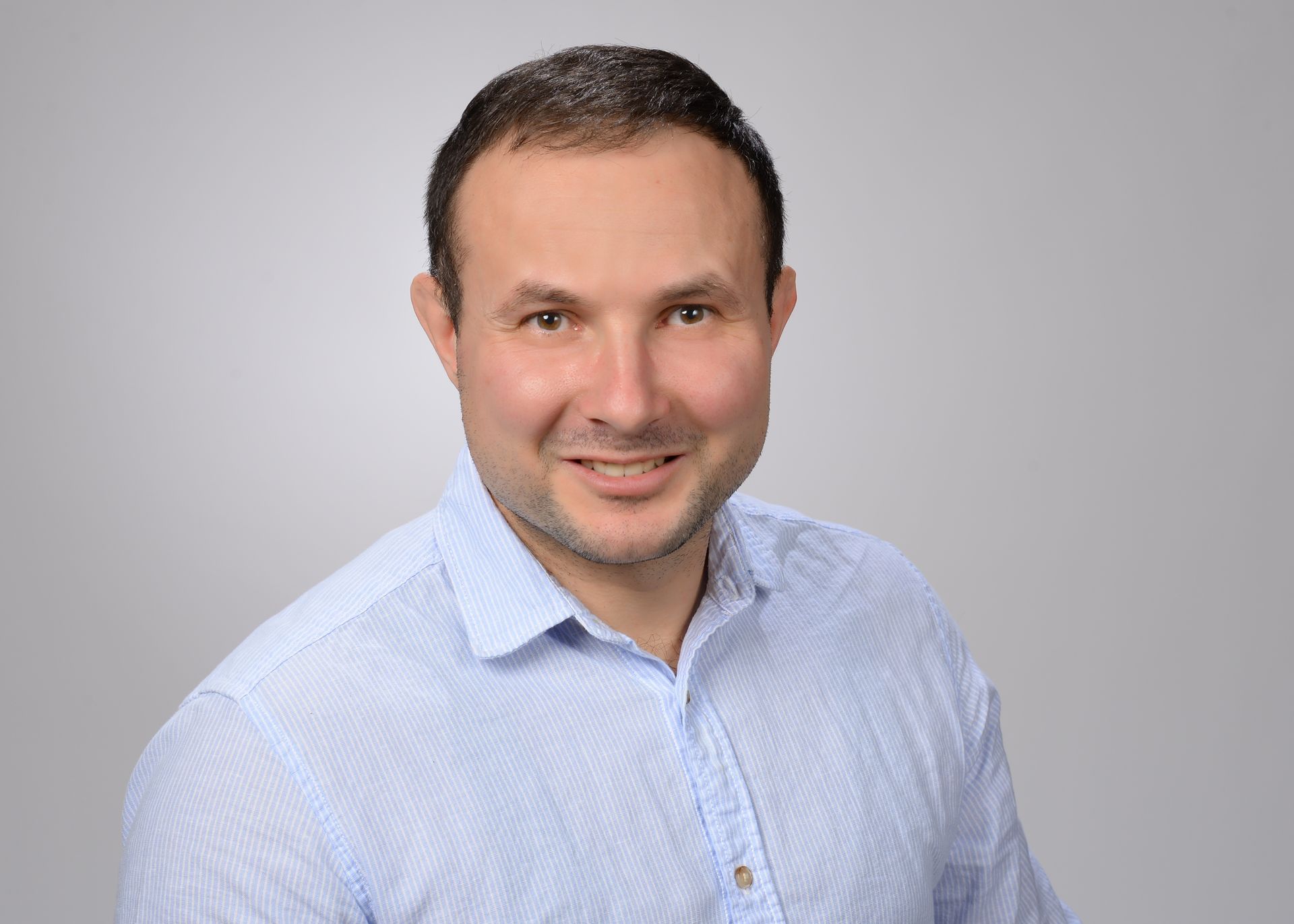 Ein Mann in einem hellblau gestreiften Hemd lächelt vor einem grauen Hintergrund in die Kamera.