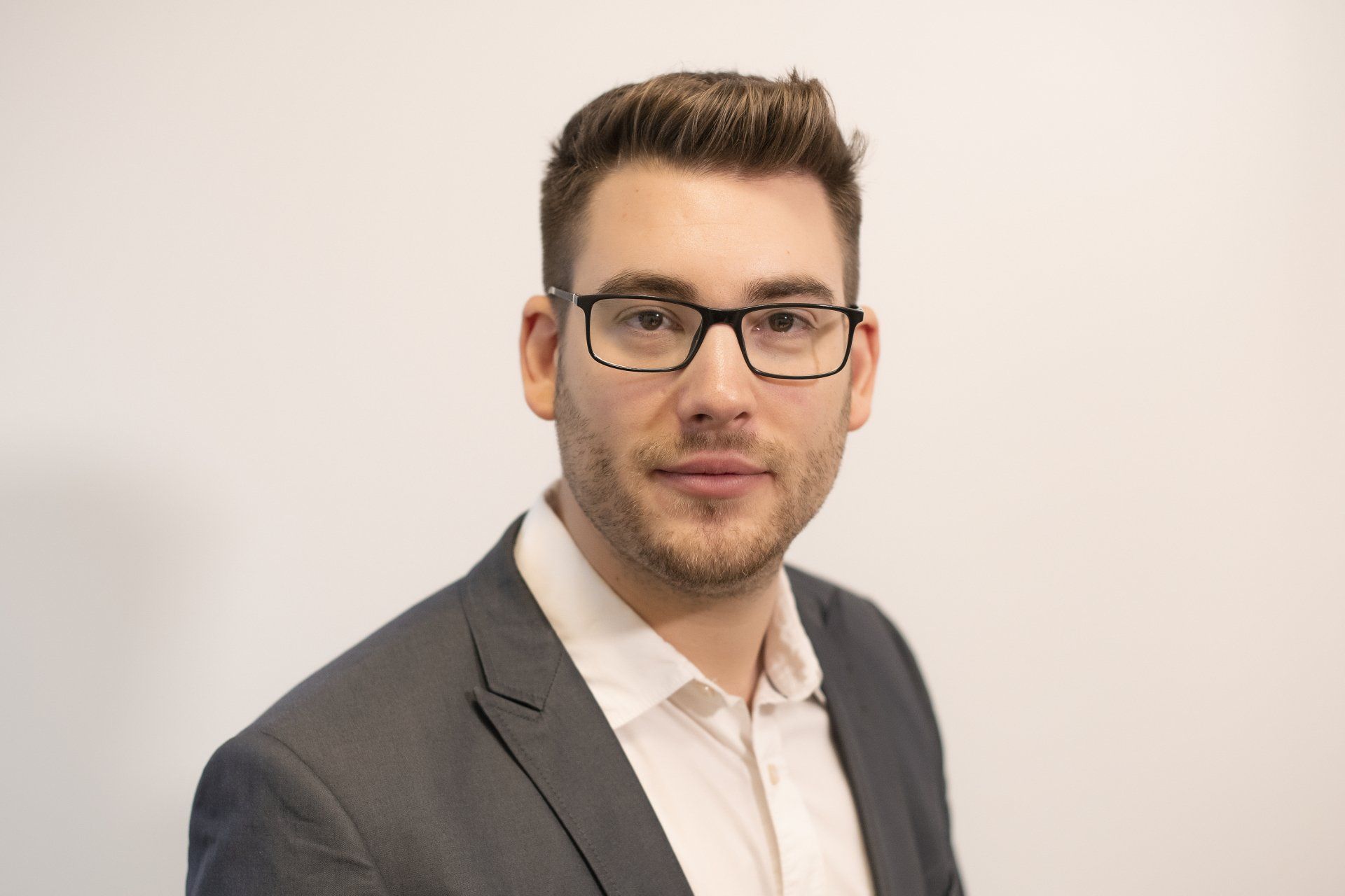 A man wearing glasses and a suit is standing in front of a white wall.