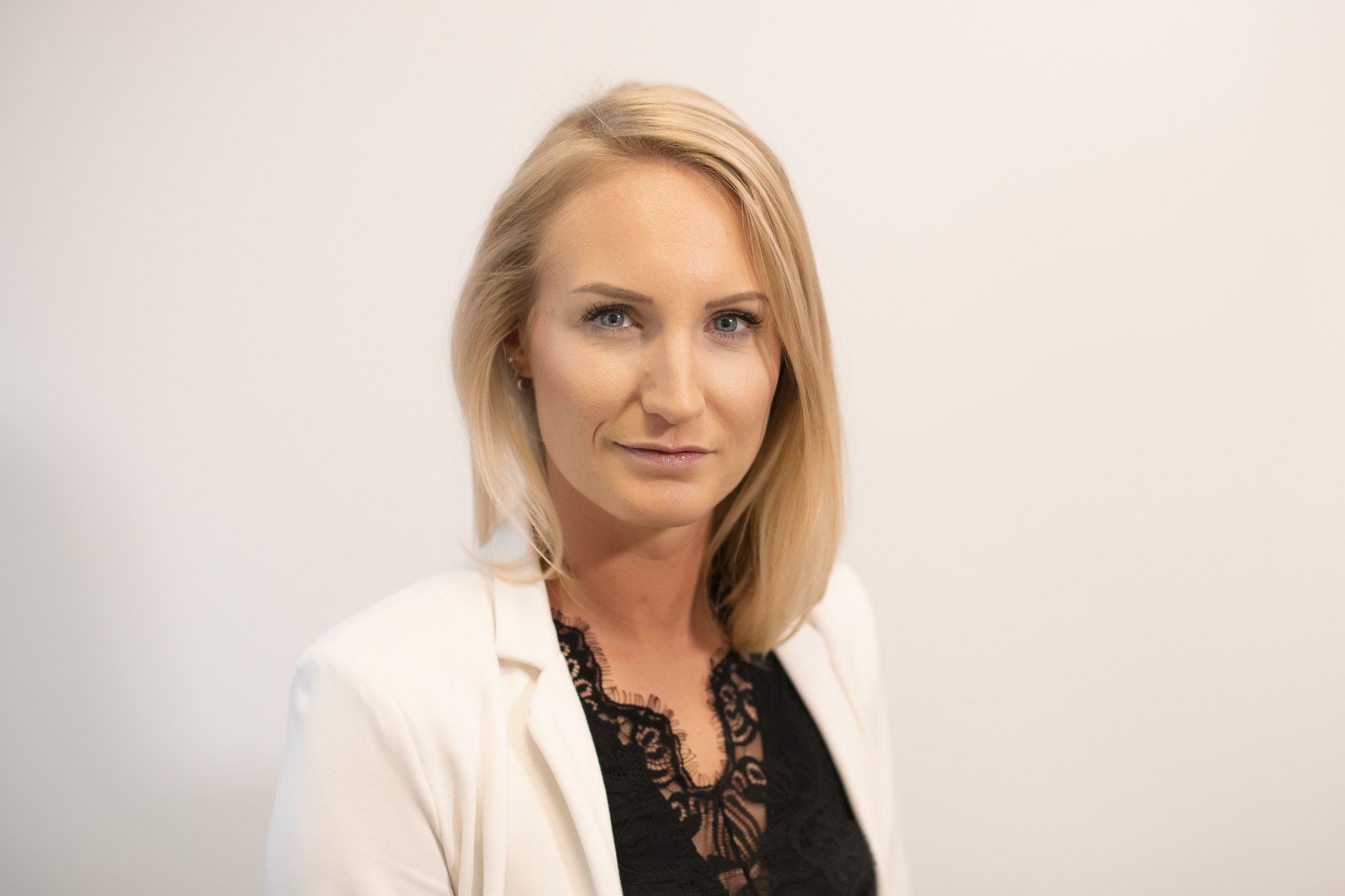 A woman in a white jacket and black top is standing in front of a white wall.