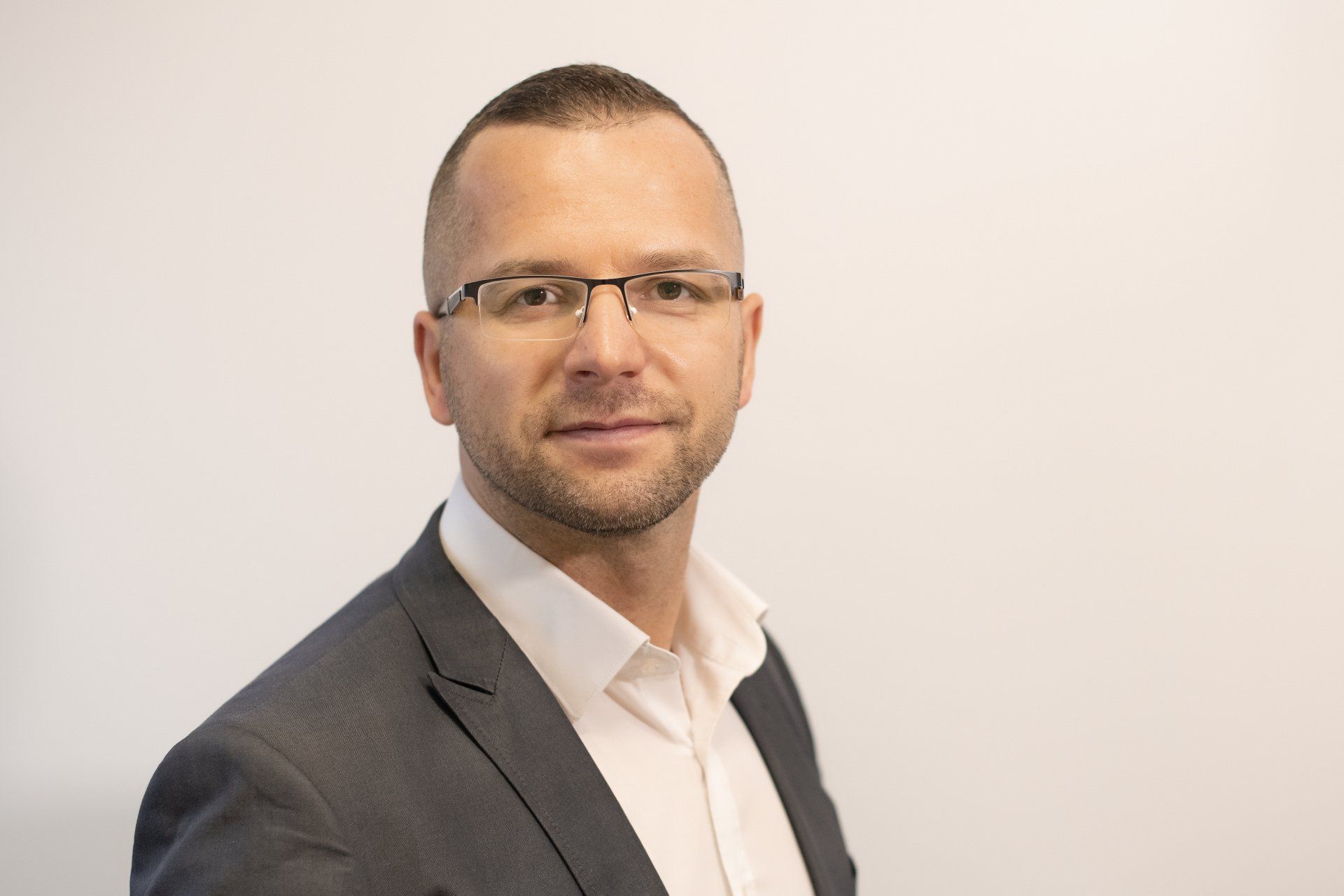 A man in a suit and glasses is standing in front of a white wall.
