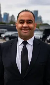 Man in black suit and tie smiles in front of vehicles and a city skyline.