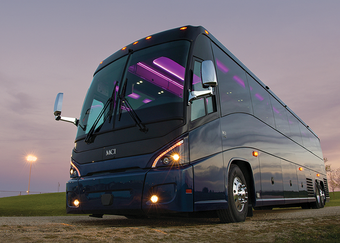 Blue MCI coach bus parked on a road, with interior purple lights illuminated. Dusk setting.