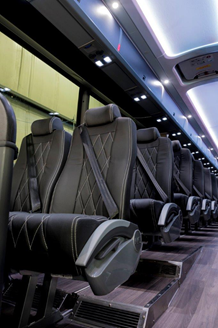 Interior view of a bus with rows of dark gray leather seats, each with a seatbelt. Overhead lights.
