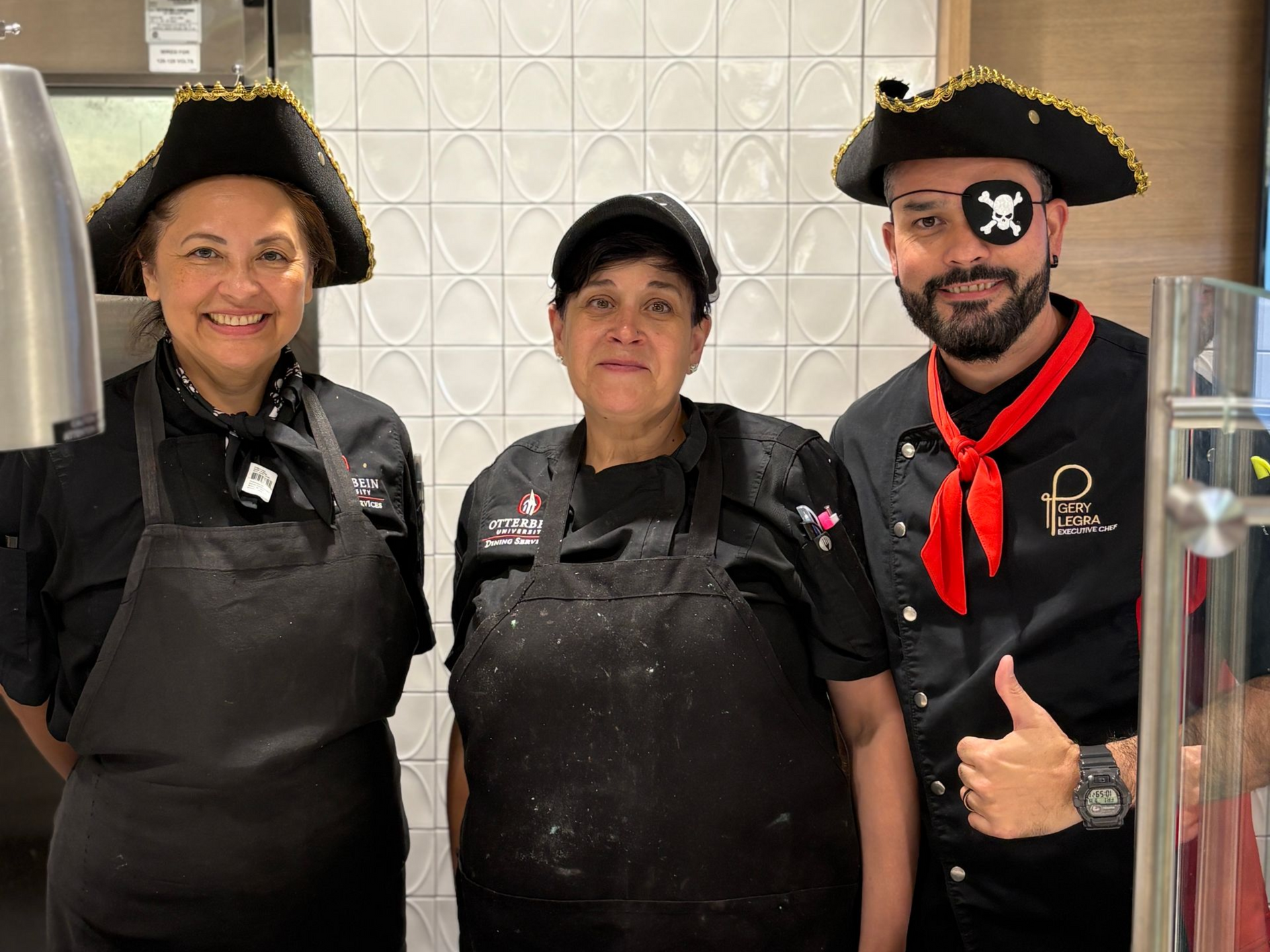 Three people in pirate hats and attire, smiling, standing in front of a white tiled wall.