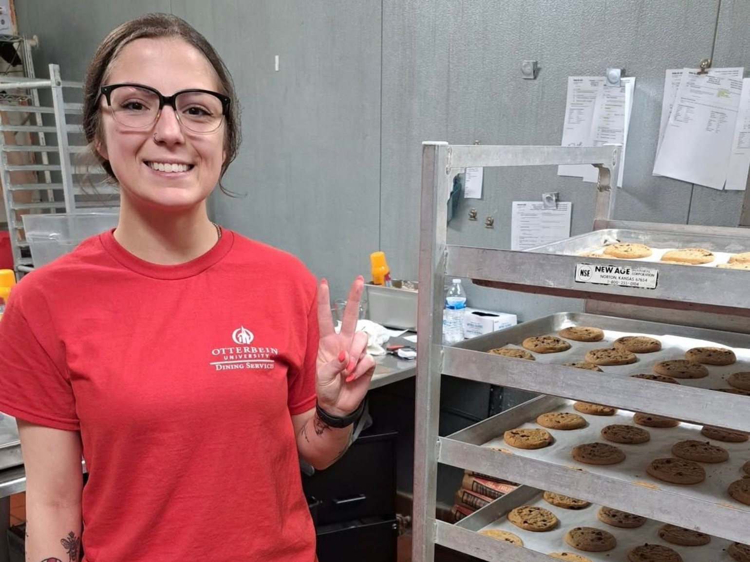 Woman in red shirt smiles, makes peace sign. Cookies on a rack in a kitchen.