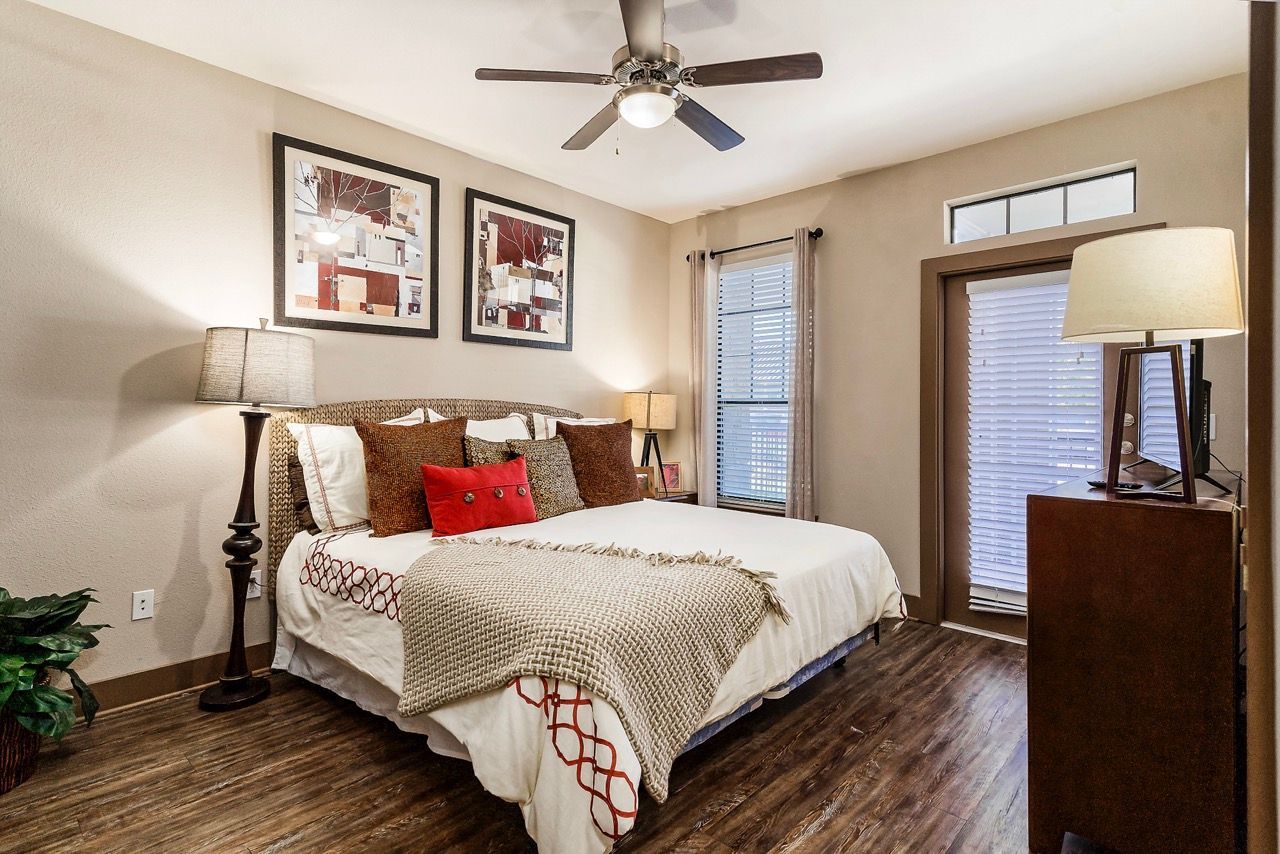 Bedroom with a king bed, multiple throw pillows, two lamps, ceiling fan, dresser, and windows with blinds.