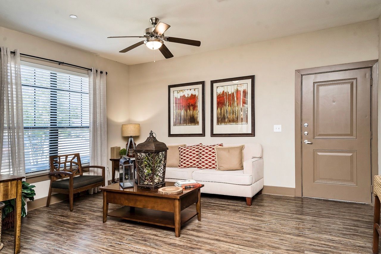 Living room of an apartment with a beige sofa, wooden coffee table, large window with blinds, and a front door.