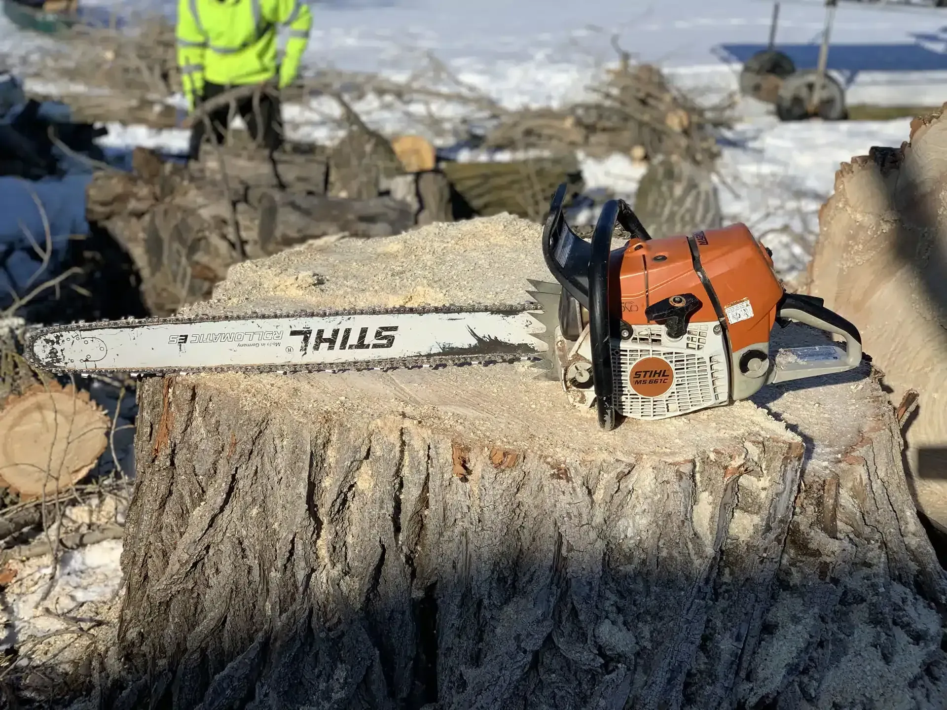 Chainsaw on a tree stump, sawdust around it; person in yellow jacket in the background.