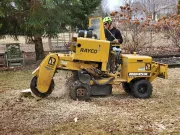 A person operating a yellow Rayco stump grinder on grass, creating wood chips.