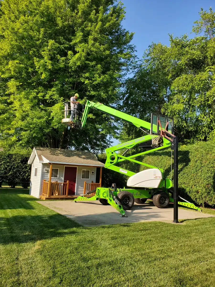 A worker in a green lift trims a tree near a playhouse in a grassy yard under a blue sky.