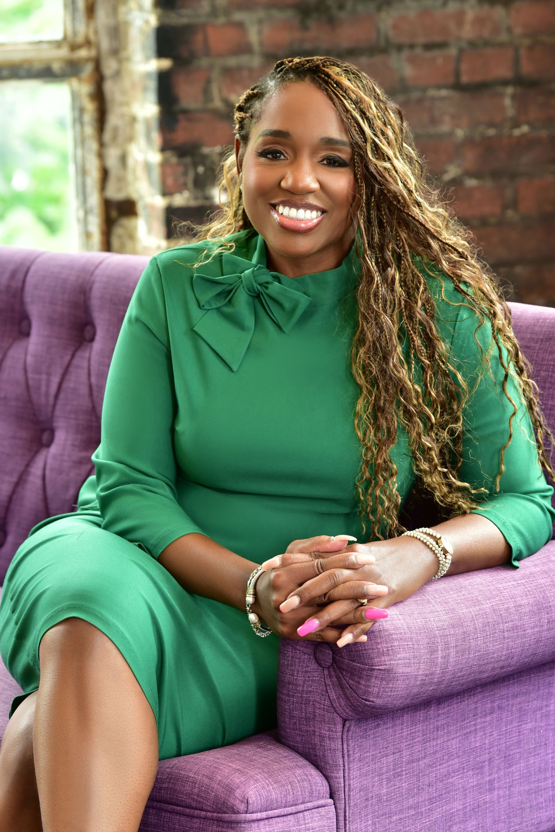 Woman in green dress sitting on purple couch, hands clasped. Braided hair, red brick wall background.