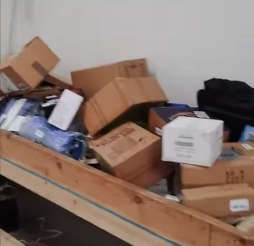 Pile of cardboard boxes and other items on a wooden surface, possibly a shelf or table, against a white wall.