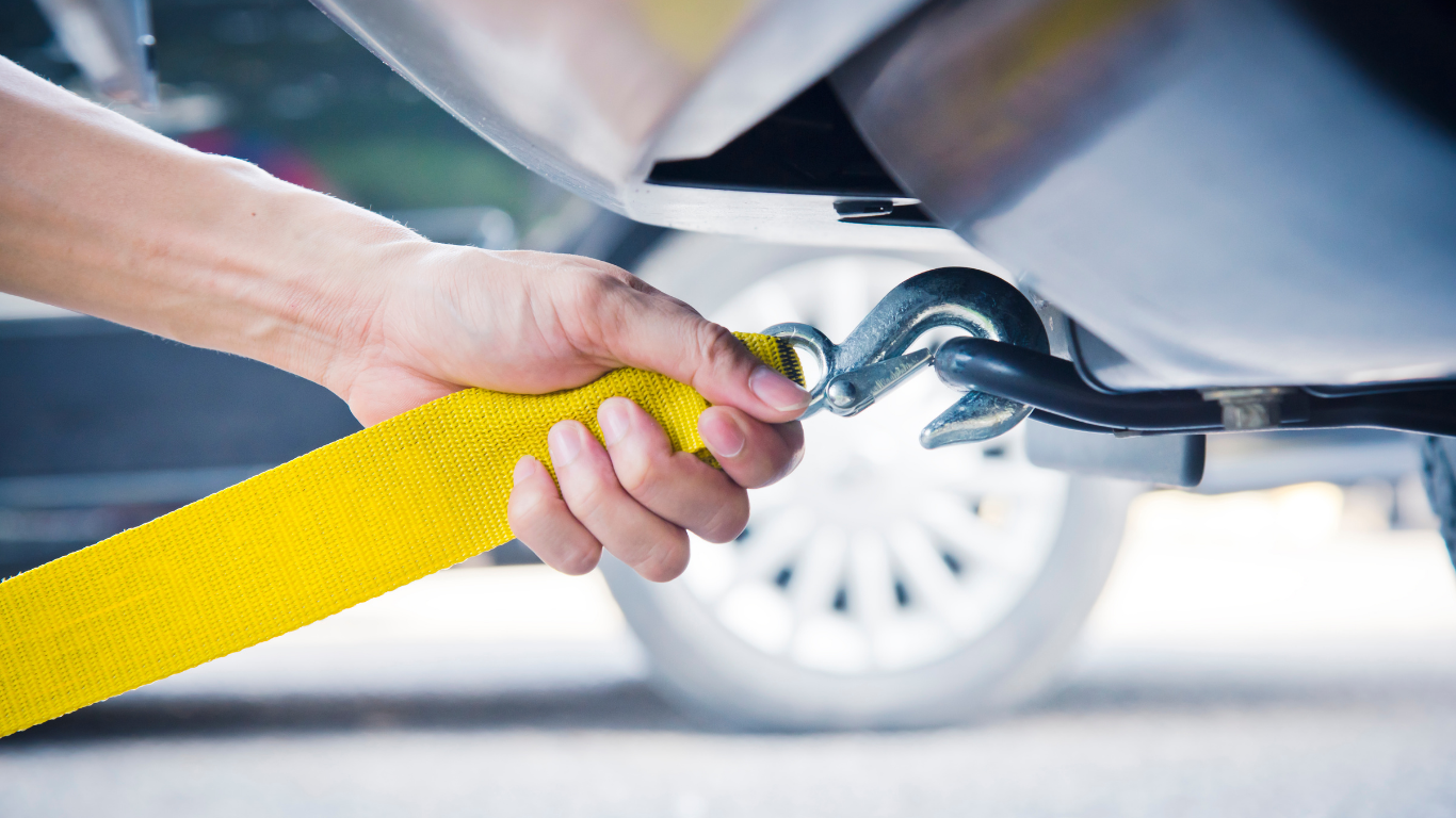 Hand connecting a yellow tow strap to a metal hook on the underside of a car.
