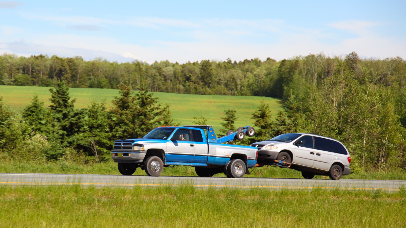Blue tow truck towing a minivan on a roadside with green fields and trees in the background.