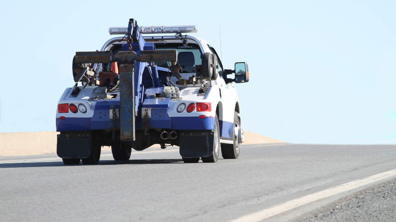 Tow truck driving away on a paved road; blue and white vehicle, sunny day.