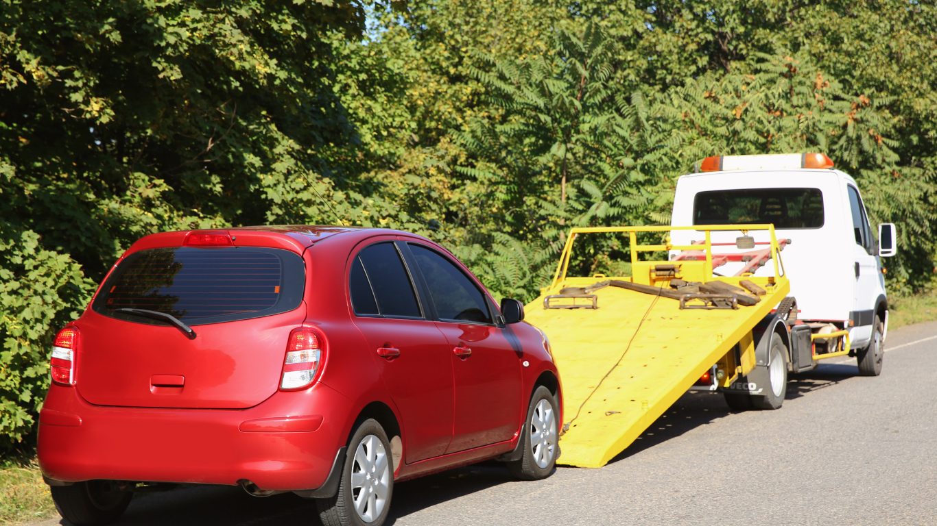 Red car being loaded onto a flatbed tow truck on a road, trees in the background.