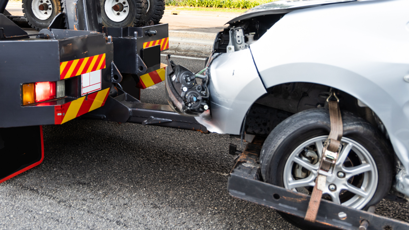Silver car with front-end damage being towed by a tow truck on a road.