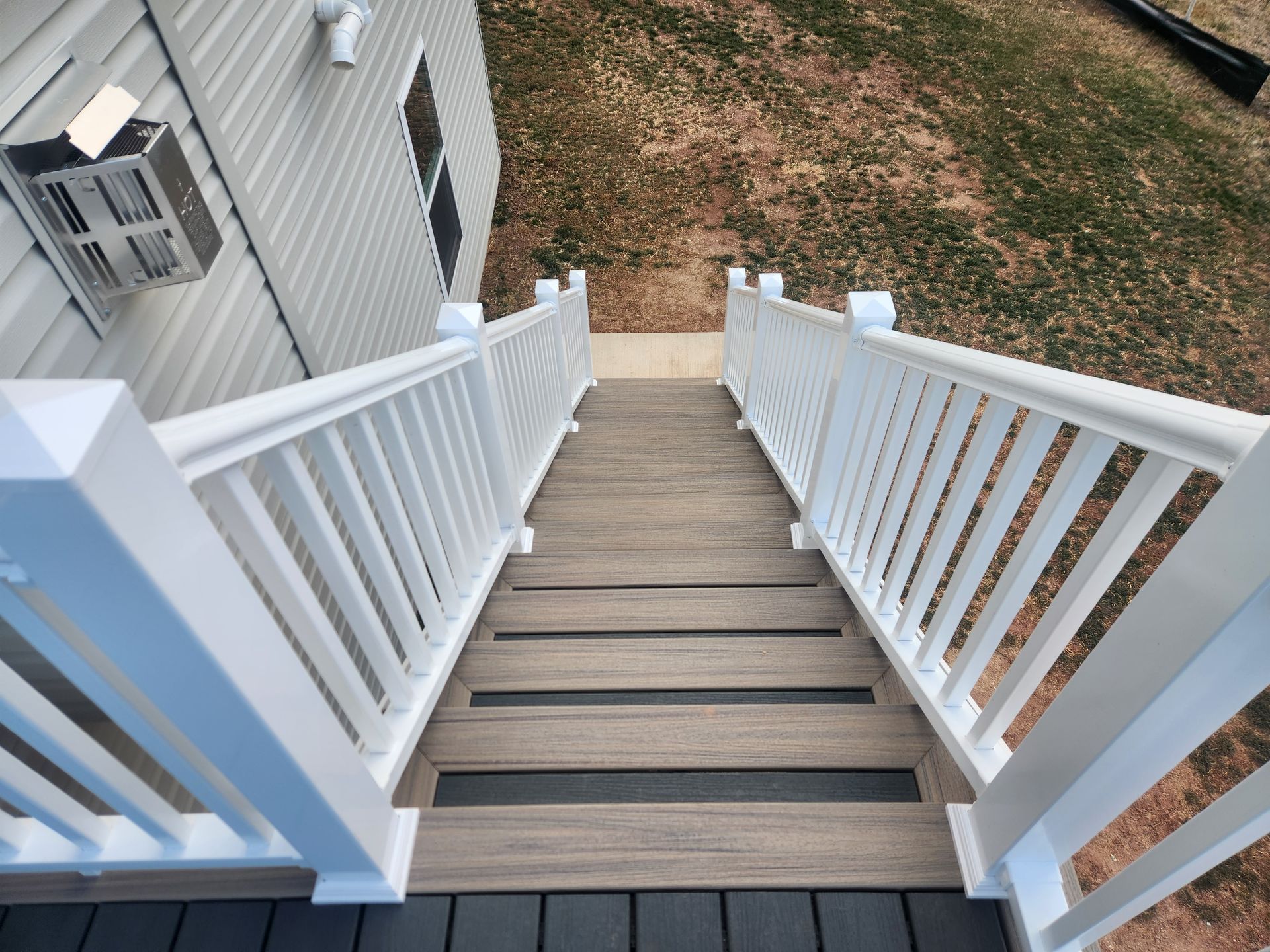 A downward view of outdoor stairs with brown wood-look steps, black anti-slip strips, and white railings, leading to a lawn.
