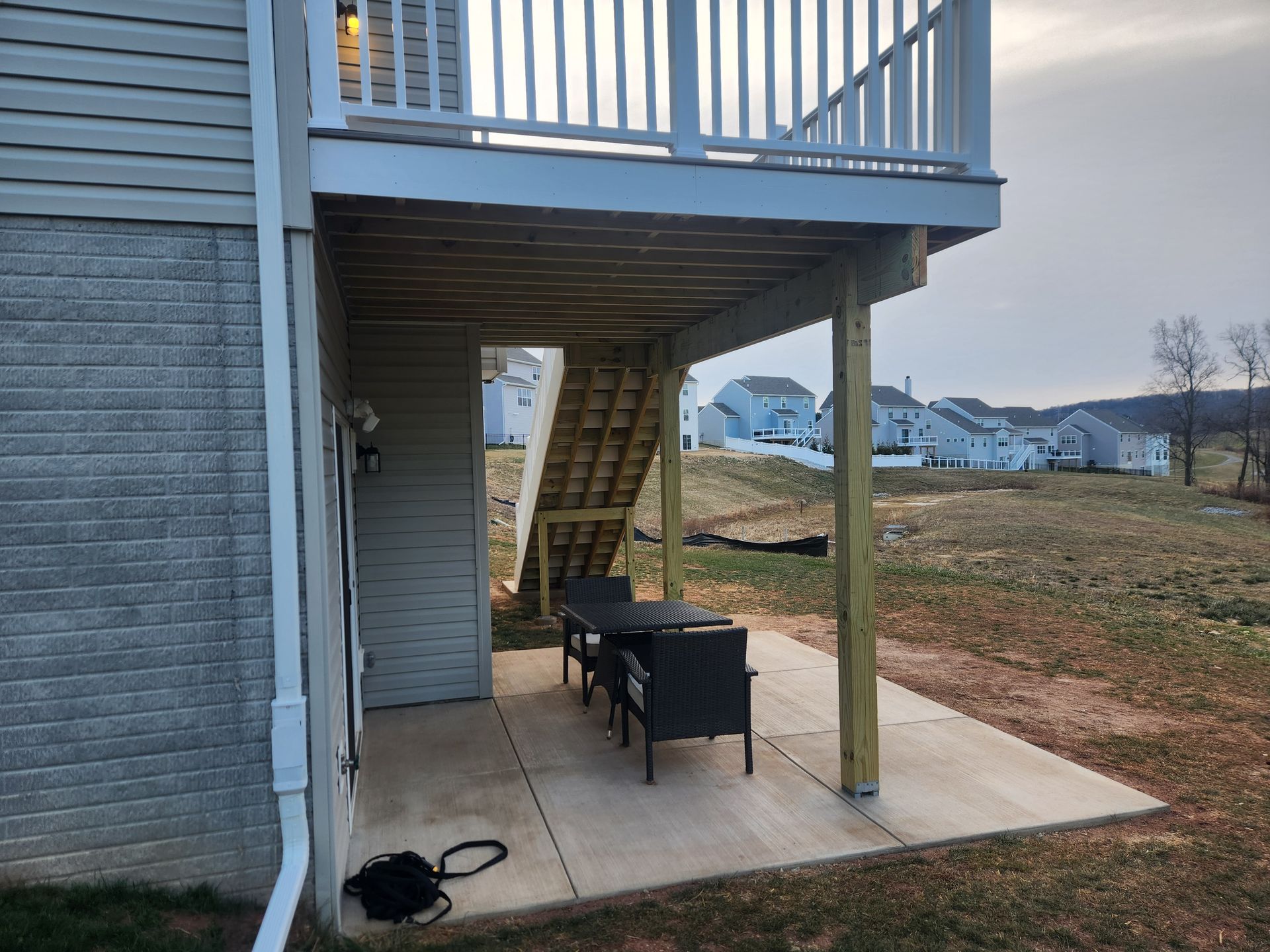 A patio beneath a wooden deck with stairs, featuring a small table, two chairs, and suburban houses in the background.
