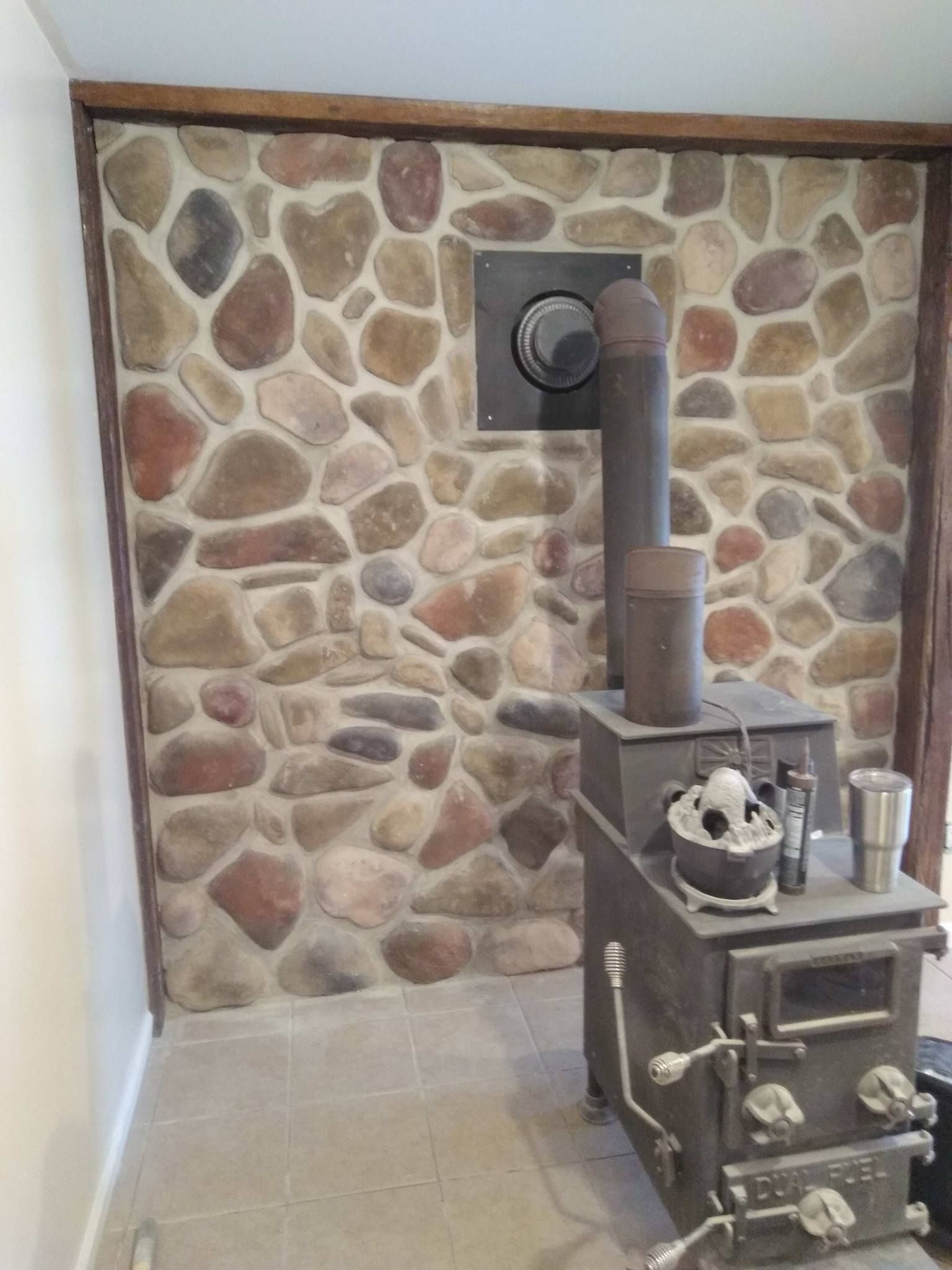 Stone-faced wall with black stove and chimney. A square metal plate holds the flue, with wooden trim.