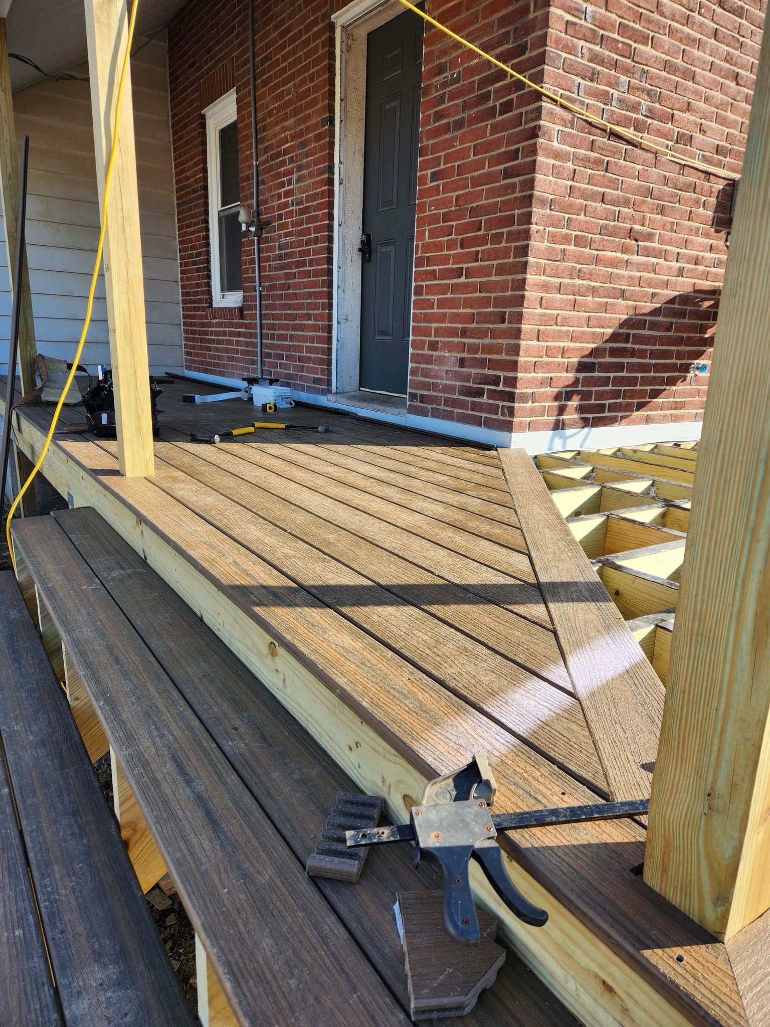 A porch under construction. New decking being installed; lumber and tools visible. Brick building background.