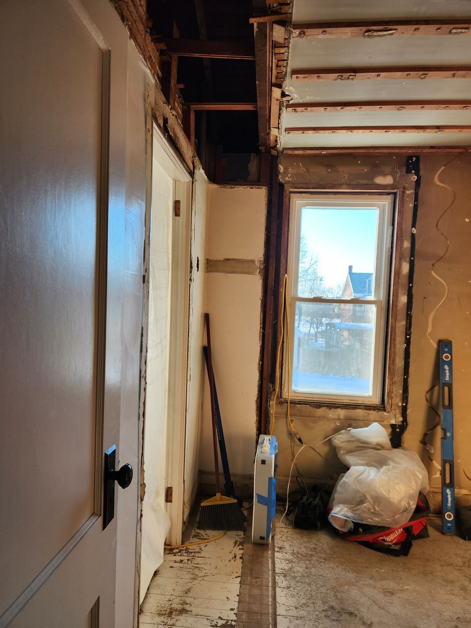Room under renovation, showing exposed wall studs, window, door, and debris on floor.