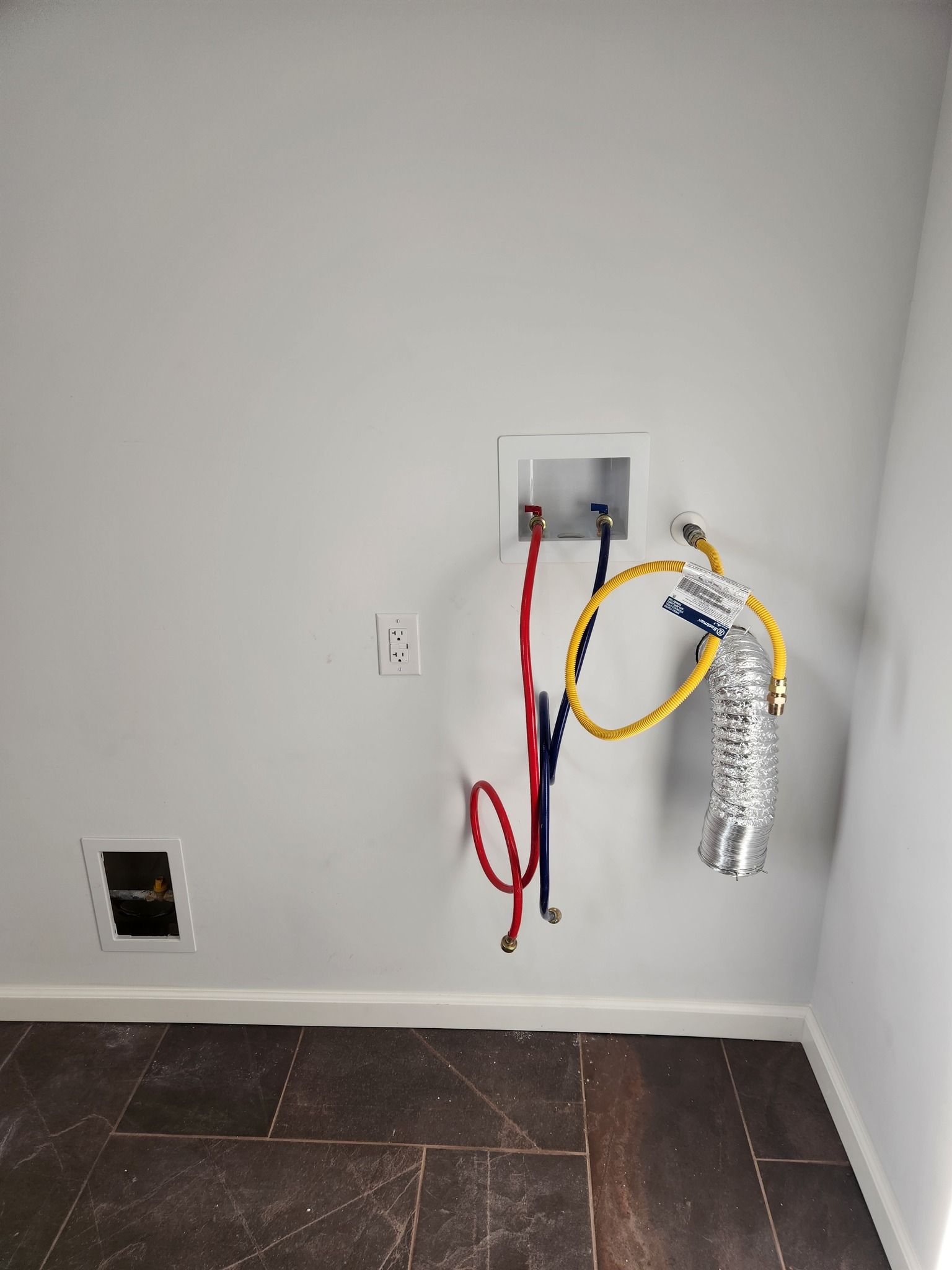White laundry room wall with water pipes, electrical outlet, and vent hose for a dryer.