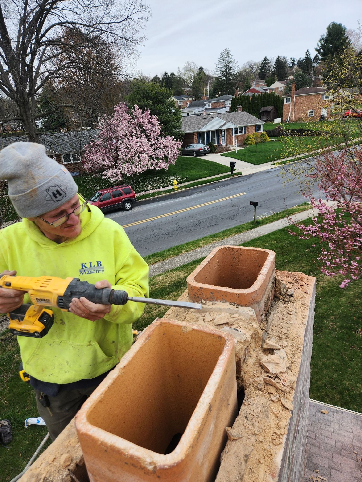 Person using a power tool on a chimney. They wear a yellow sweatshirt and beanie. Houses and trees are in the background.